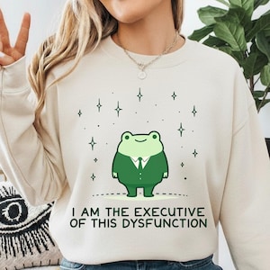 Peut inclure: Sweat-shirt crème avec une grenouille de dessin animé en costume vert et le texte "I AM THE EXECUTIVE OF THIS DYSFUNCTION." Le motif comprend des étoiles. Le sweat-shirt est de style décontracté.