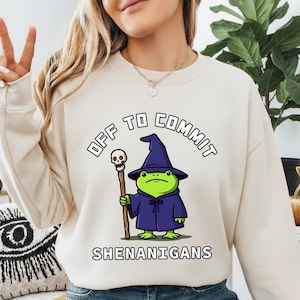 Może przedstawiać: Kremowa bluza z napisem "OFF TO COMMIT SHENANIGANS". Grafika przedstawia kreskówkową żabę w kapeluszu i szacie czarodzieja, trzymającą laskę z czaszką. Żaba jest zielona, szata fioletowa, a laska brązowa.