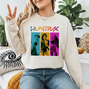 May include: A cream-colored sweatshirt with the word "HUNTRX" in a colorful, stylized font. The graphic features three women in colorful panels. The woman on the left is in a blue panel, the middle woman is in a yellow panel, and the woman on the right is in a pink panel.