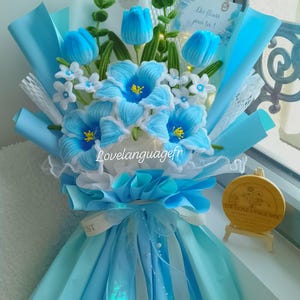Op de afbeelding: Een boeket van blauwe en witte stoffen bloemen, waaronder tulpen en grotere bloemen met gele centra. Het boeket is verpakt in lichtblauw papier en lint, met een label.