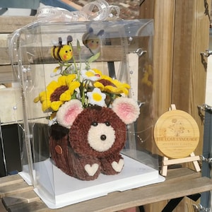Peut inclure: Composition florale en forme d'ours en peluche fait à la main, dans une boîte transparente. L'ours est brun avec des oreilles roses et des pieds en forme de cœur blancs. L'arrangement comprend des tournesols jaunes, des marguerites blanches et deux décorations d'abeilles. Un petit panneau en bois indique "THE LOVE LANGUAGE".