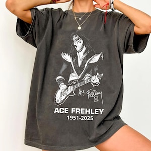 Op de afbeelding: Donkergrijs t-shirt met een witte afbeelding van Ace Frehley die gitaar speelt. De tekst "ACE FREHLEY 1951-2025" staat onder de afbeelding. Het shirt heeft korte mouwen en een relaxte pasvorm.
