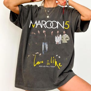 May include: Dark gray t-shirt featuring the band Maroon 5. The shirt displays the band's name in white and yellow, with a photo of the band members. Below the photo, the text "Love is like" and "2025 US FALL TOUR" are in yellow.