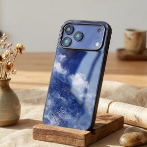 Może przedstawiać: Etui na smartfon z motywem błękitnego nieba i chmur. Etui ma czarną obwódkę i przezroczyste, błyszczące wykończenie. Etui na telefon jest prezentowane na drewnianym stojaku.