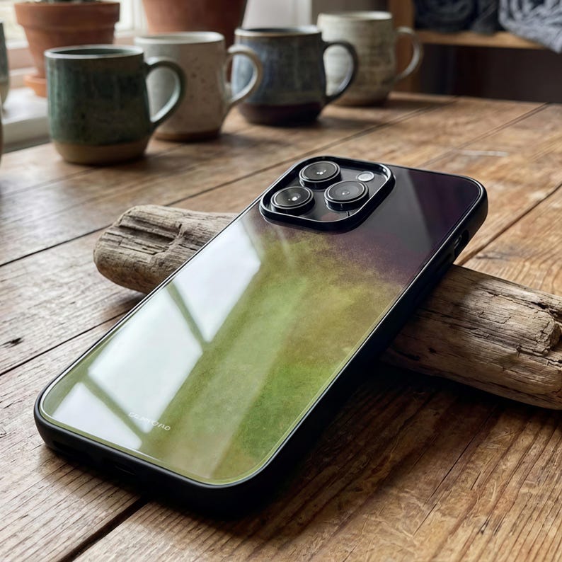 May include: A black iPhone case with a gradient design transitioning from green to purple. The case has a glossy finish and a black border. The phone's camera lenses are visible. The case is resting on a piece of driftwood.