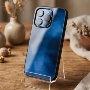 Könnte beinhalten: Ein blaues Smartphone mit schwarzem Rahmen wird auf einem transparenten Acrylständer präsentiert. Das Telefon hat mehrere Kameralinsen. Der Text "DUSTY STONE" ist unten auf dem Telefon sichtbar. Der Hintergrund umfasst eine Holzoberfläche und dekorative Elemente.