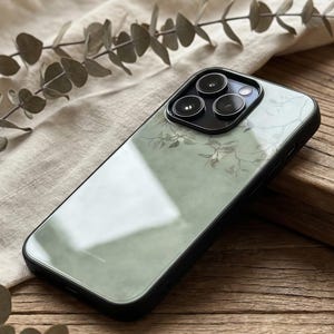 以下が含まれることがあります： 黒い携帯電話ケース、フローラルデザイン。 ケースは、くすんだ緑色の背景と、枝と葉の繊細なパターンを備えた透明で光沢のある背面を備えています。 黒い縁とカメラレンズプロテクターが付いています。
