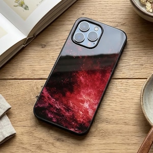 May include: A smartphone case with a black frame and a vibrant red and black abstract design. The case has a glossy finish and features a camera module with three lenses. The phone case is on a wooden surface.