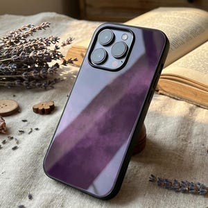 May include: A glossy, purple-gradient phone case with a black border. The case features a camera module with three lenses and a flash. The case is angled, revealing its reflective surface. The background includes dried lavender and an open book.