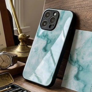 Op de afbeelding: Een smartphonehoesje met een glanzend, lichtgroen en wit marmerpatroon en een zwarte cameramodule met drie lenzen. Het hoesje heeft een zwart frame en de tekst TOY STUNO in kleine witte letters bij de onderste rand gedrukt.