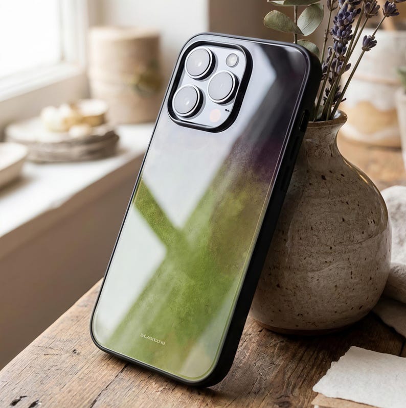 May include: A black iPhone case with a green and purple watercolor design. The case has a glossy finish and a black border. The case is leaning against a vase with lavender sprigs.