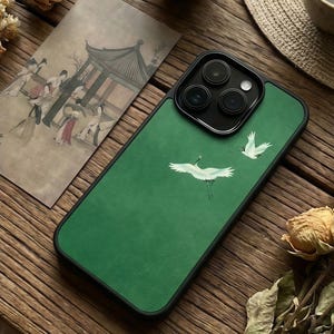 May include: Green phone case with a black border, featuring a white crane design. The case is next to a vintage illustration of people in traditional clothing and a dried flower on a wooden surface.