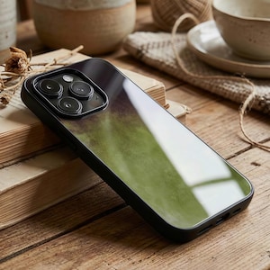 May include: A black phone case with a green and purple watercolor design. The case has a glossy finish and a black border. The phone case is resting on a wooden surface next to a book.