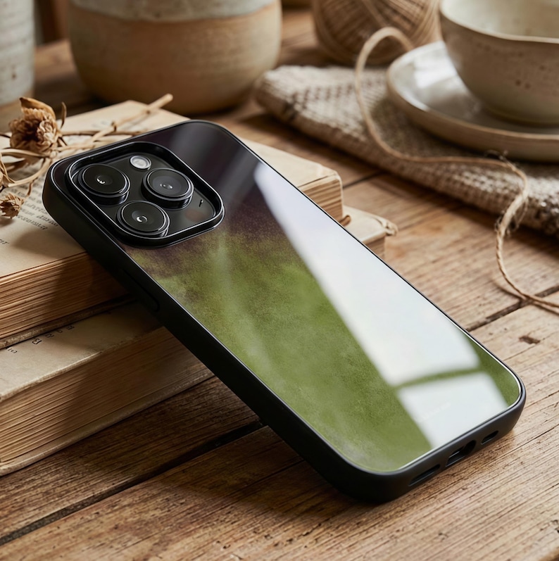 May include: A black phone case with a green and purple watercolor design. The case has a glossy finish and a black border. The phone case is resting on a wooden surface next to a book.