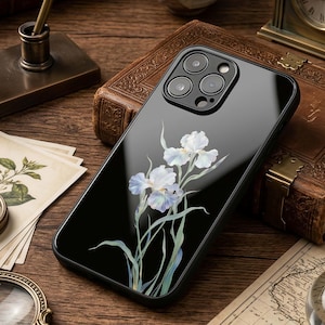May include: A black smartphone case featuring a botanical illustration of white and pale blue iris flowers with green stems and leaves. The case has a glossy finish and a cutout for a triple-lens camera system on the upper left corner.
