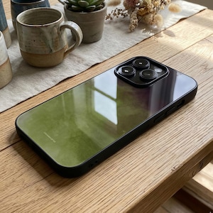 May include: A black phone case with a green and purple gradient design. The case has a glossy finish and a black border. The camera lenses are visible at the top. The case is on a wooden surface.