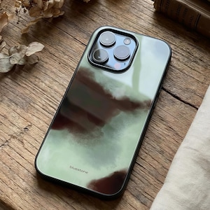 Könnte beinhalten: Eine Smartphone-Hülle mit einem grün-braunen Aquarelldesign. Die Hülle hat einen schwarzen Rand und das Wort "bluestone" ist unten aufgedruckt. Die Hülle hat eine glänzende Oberfläche und bedeckt die Rückseite und die Seiten des Telefons.