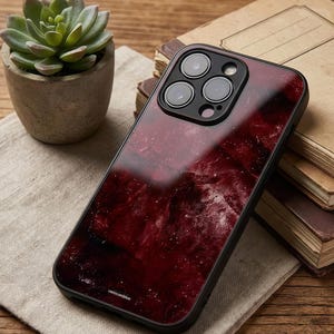 以下が含まれることがあります： 黒いスマートフォンケース。深紅の大理石模様のデザイン。ケースは光沢仕上げで、黒い縁取り。カメラレンズが上部に表示されています。「omncomblue」の文字が下部に印刷されています。ケースはベージュ色の布の上に置かれています。