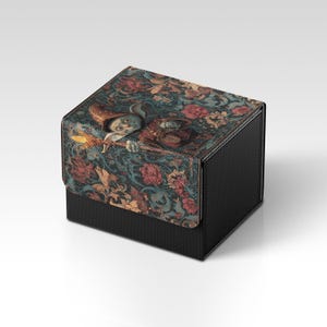 May include: A black box with a textured surface and a patterned lid. The lid features a whimsical illustration of a figure holding a glowing object, set against a backdrop of floral and foliate designs in shades of blue, red, and gold.