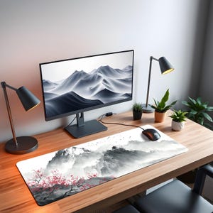 Puede incluir: Una configuraci&oacute;n de escritorio con un monitor de computadora que muestra una escena de monta&ntilde;a en escala de grises, un rat&oacute;n negro y una alfombrilla de rat&oacute;n larga con un dise&ntilde;o de monta&ntilde;a y flores de cerezo. Tambi&eacute;n se ven dos l&aacute;mparas de escritorio negras y plantas en macetas.