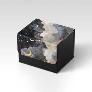 May include: A black box with a lid displaying a night sky design. The lid features gold stars, crescent moons, and clouds in blue, grey, and gold. The box has a textured, dark surface. The design evokes a dreamy, celestial theme.