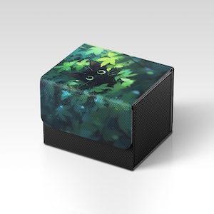 May include: A black box with a lid featuring a cat peeking through green leaves. The cat has green eyes. The box has a black textured exterior.