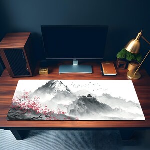 Puede incluir: Alfombrilla de rat&oacute;n rectangular con un paisaje asi&aacute;tico sereno. El dise&ntilde;o muestra monta&ntilde;as brumosas, una pagoda y flores de cerezo en tonos grises, negros y rojos. La alfombrilla est&aacute; sobre un escritorio de madera con un monitor.