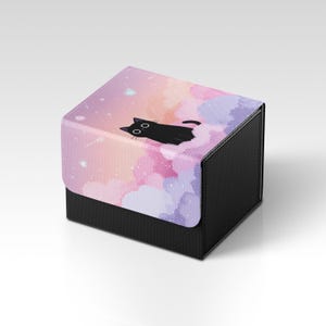 Puede incluir: Una caja negra y gris con una tapa que presenta un diseño caprichoso. La tapa muestra un gato negro descansando sobre nubes rosas, moradas y naranjas con estrellas. La caja tiene una apariencia texturizada.
