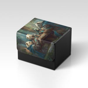 Könnte beinhalten: Eine schwarze, strukturierte Kartenaufbewahrungsbox mit einem Klappdeckel. Der Deckel zeigt eine Fantasy-Kunstszene mit Figuren in Blau- und Brauntönen. Die Box ist für die Aufbewahrung von Sammelkarten oder anderen Sammlerstücken konzipiert.