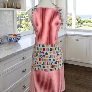 May include: A kitchen apron on a mannequin featuring a red and white vertical striped pattern on the top and bottom sections. The center panel displays a white background with repeating colorful text reading Merry, Bright, Happy, and Jolly, interspersed with yellow stars and green pine tree icons.