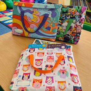 May include: Three colorful tote bags with unique designs sit on a wooden table. One bag features a vibrant, psychedelic pattern, another has a cartoon monster design, and the third displays an owl pattern. Books are visible inside the bags.