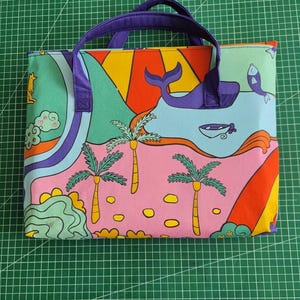 May include: A rectangular fabric tote bag with purple handles featuring a colorful graphic print of a landscape with a whale, palm trees, a sun, and a fish. The design includes vibrant sections of pink, orange, yellow, green, and blue colors.