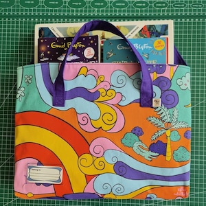 May include: A rectangular fabric tote bag with purple handles and a colorful pattern featuring a rainbow, stylized clouds, a palm tree, and a small alligator. A white label with the text 'I belong to:' is stitched on the bottom left corner.