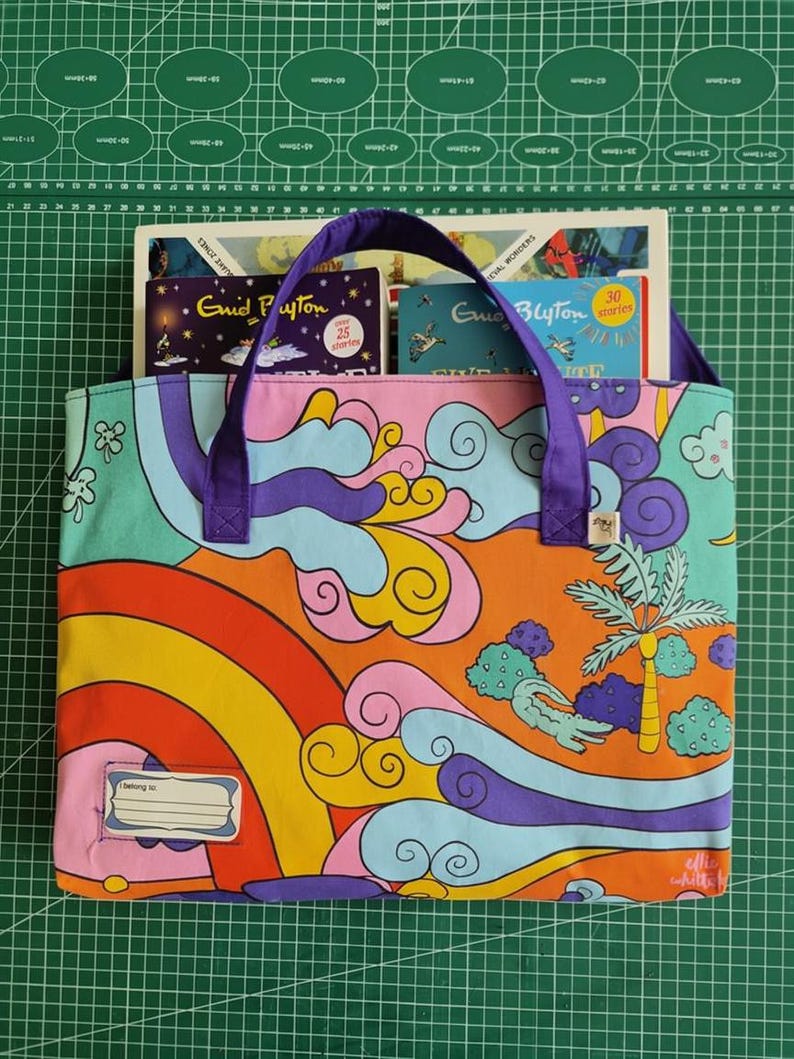 May include: A rectangular fabric tote bag with purple handles and a colorful pattern featuring a rainbow, stylized clouds, a palm tree, and a small alligator. A white label with the text 'I belong to:' is stitched on the bottom left corner.