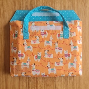 May include: A fabric bag with a bright orange background featuring a repeating pattern of white llamas wearing colorful blankets and garlands. The bag has a light blue handle and a matching light blue flap with a white star pattern and velcro closure.