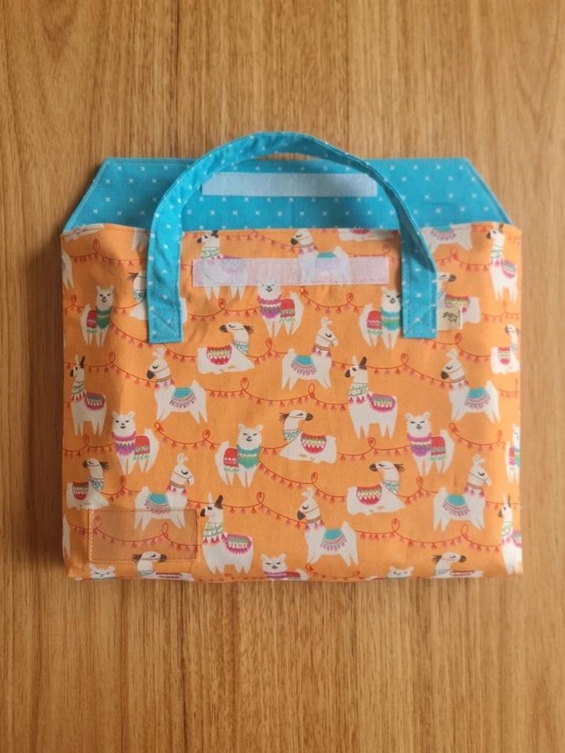 May include: A fabric bag with a bright orange background featuring a repeating pattern of white llamas wearing colorful blankets and garlands. The bag has a light blue handle and a matching light blue flap with a white star pattern and velcro closure.