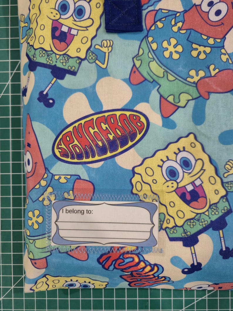 Pu&ograve; includere: Una custodia in tessuto con un motivo ripetuto di SpongeBob SquarePants e Patrick Stella su uno sfondo azzurro con forme di fiori bianchi. Un'etichetta rettangolare bianca con il testo I belong to: &egrave; cucita sulla parte anteriore.