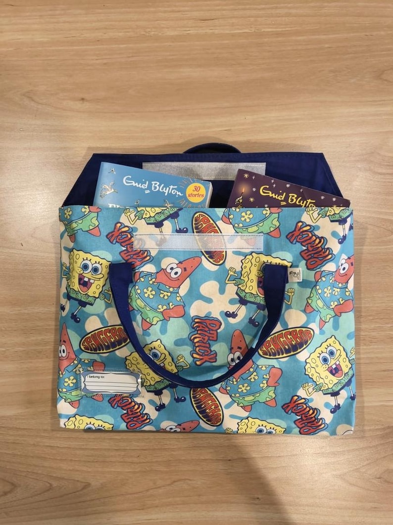 Pu&ograve; includere: Una borsa in tessuto blu per libri con un motivo ripetuto dei personaggi SpongeBob SquarePants e Patrick Stella. La borsa presenta manici blu scuro, una chiusura a strappo e un'etichetta bianca con la scritta "Belong to" stampata sul davanti.