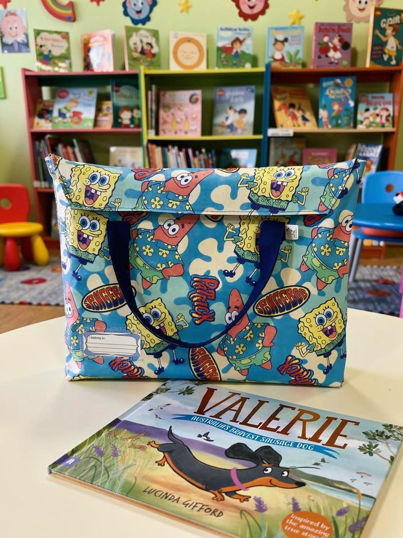 Blue SpongeBob SquarePants tote bag / library book bag with SpongeBob, Patrick and cartoon characters print, blue handles and name tag – shown in children’s library with the book of Valerie the dachshund