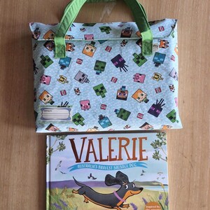 Pu&ograve; includere: Una borsa in tessuto azzurro con manici verdi caratterizzata da un motivo ripetuto di personaggi pixelati e blocchi TNT, con un'etichetta per il nome. Sotto c'&egrave; un libro con copertina rigida intitolato Valerie: Australia's Bravest Sausage Dog.