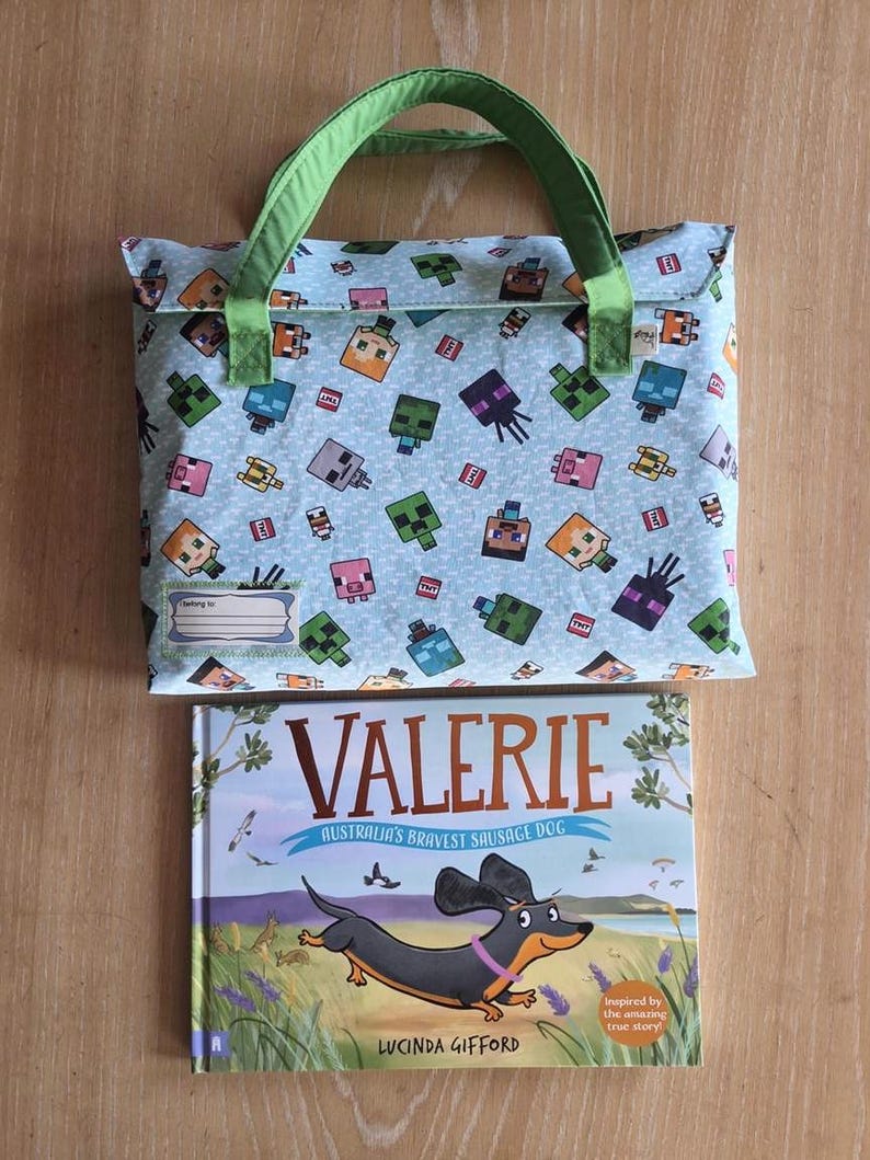 Pu&ograve; includere: Una borsa in tessuto azzurro con manici verdi caratterizzata da un motivo ripetuto di personaggi pixelati e blocchi TNT, con un'etichetta per il nome. Sotto c'&egrave; un libro con copertina rigida intitolato Valerie: Australia's Bravest Sausage Dog.