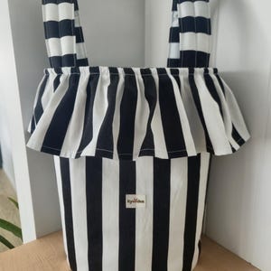 May include: A cylindrical fabric storage basket featuring a black and white vertical striped pattern. The basket has two matching striped handles and a ruffled fabric trim around the top edge. A small rectangular label reads kye&dee.
