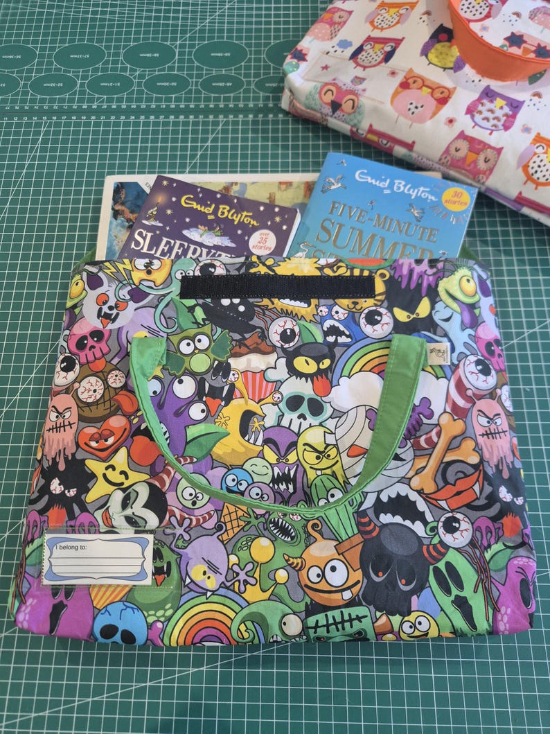 May include: A rectangular fabric tote bag featuring a colorful, dense pattern of cartoon monsters, skulls, eyeballs, and rainbows. The bag has light green fabric handles, a black velcro closure, and a clear plastic name tag label reading I belong to.