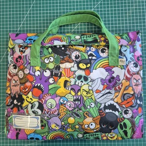 May include: A rectangular fabric bag with light green handles featuring a colorful all-over print of cartoon monsters, skulls, rainbows, and bones. A clear plastic name tag holder is attached to the bottom left corner with the text I belong to.