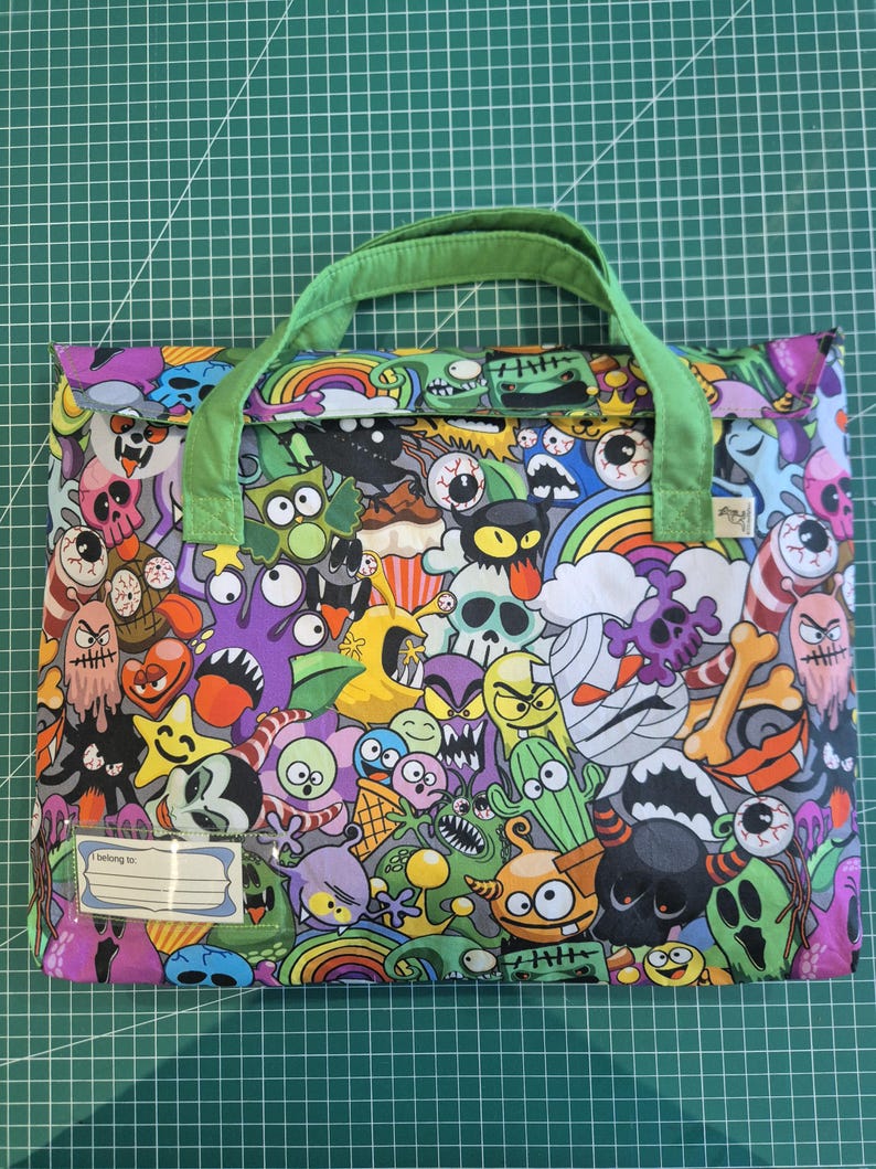 May include: A rectangular fabric bag with light green handles featuring a colorful all-over print of cartoon monsters, skulls, rainbows, and bones. A clear plastic name tag holder is attached to the bottom left corner with the text I belong to.