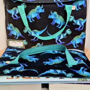 Kids dinosaur library bag tote bag dark blue background with blue/green Triceratops and Tyrannosaurus rex aka T.rex pattern, turquoise teal handles, name label “I Belong To,” perfect book bag for children, displayed on stack of kids’ nonfiction books, including What’s Where in the World.