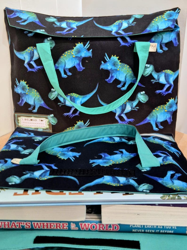 Kids dinosaur library bag tote bag dark blue background with blue/green Triceratops and Tyrannosaurus rex aka T.rex pattern, turquoise teal handles, name label “I Belong To,” perfect book bag for children, displayed on stack of kids’ nonfiction books, including What’s Where in the World.