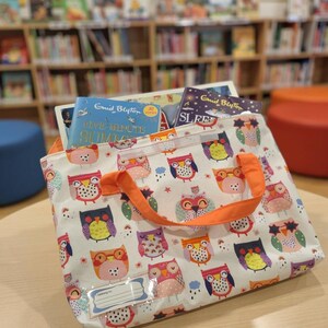 May include: A white fabric tote bag featuring a repeating pattern of colorful owls in shades of orange, pink, purple, and blue. The bag has bright orange handles and a clear plastic name tag holder on the front. Two books are visible inside the bag.