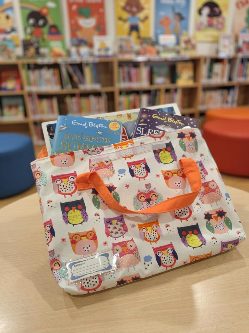 May include: A white fabric tote bag featuring a repeating pattern of colorful owls in shades of orange, pink, purple, and blue. The bag has bright orange handles and a clear plastic name tag holder on the front. Two books are visible inside the bag.