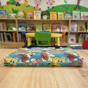 Pu&ograve; includere: Un oggetto rettangolare, piatto e rivestito in tessuto con una vivace stampa all-over di SpongeBob SquarePants e Patrick Stella su sfondo azzurro. L'oggetto presenta manici in tessuto blu scuro e una tasca in plastica trasparente per etichette.
